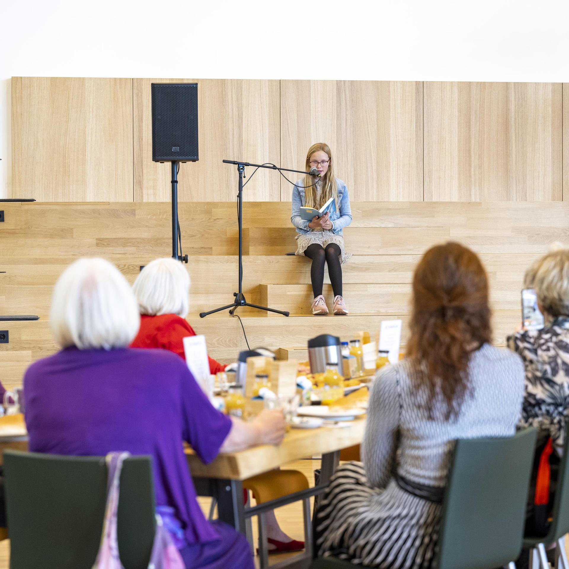 Elderly people are read to by a girl during lunch at De Nieuwe Kolk
